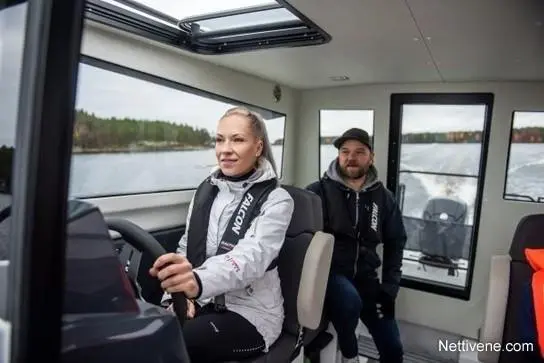 Woman steering a motorboat, wearing a life jacket, with a smiling man seated next to her; scenic water view outside.